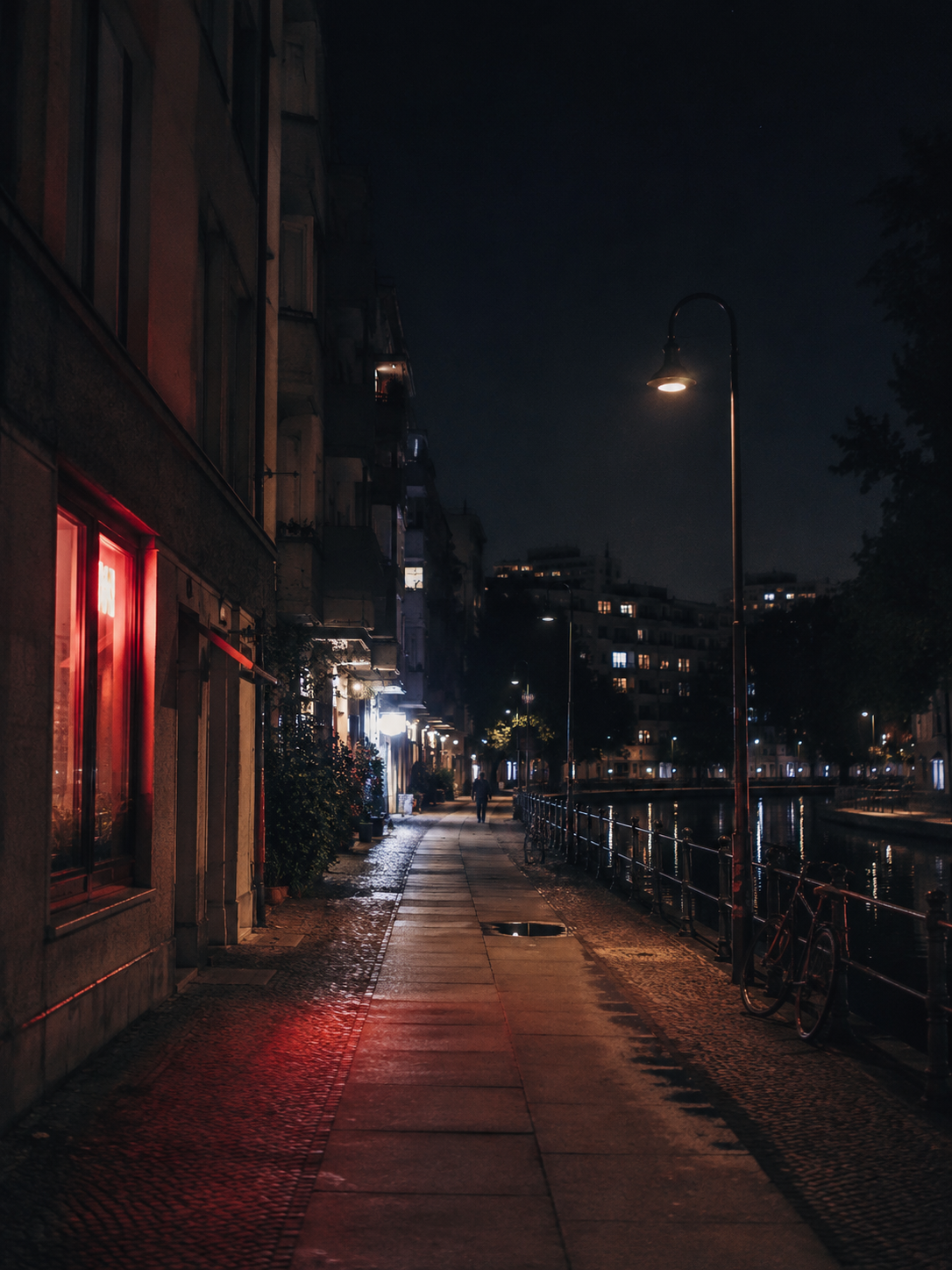 Berlin summer night by the canal with wet pavement, red neon reflections, apartment buildings, bicycles and soft city lights.